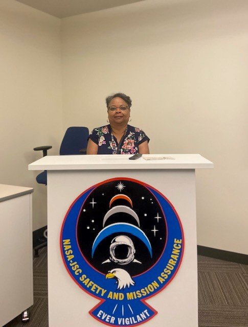A woman sits behind a white podium with a large patch showing planets, an eagle, and an astronaut helmet.